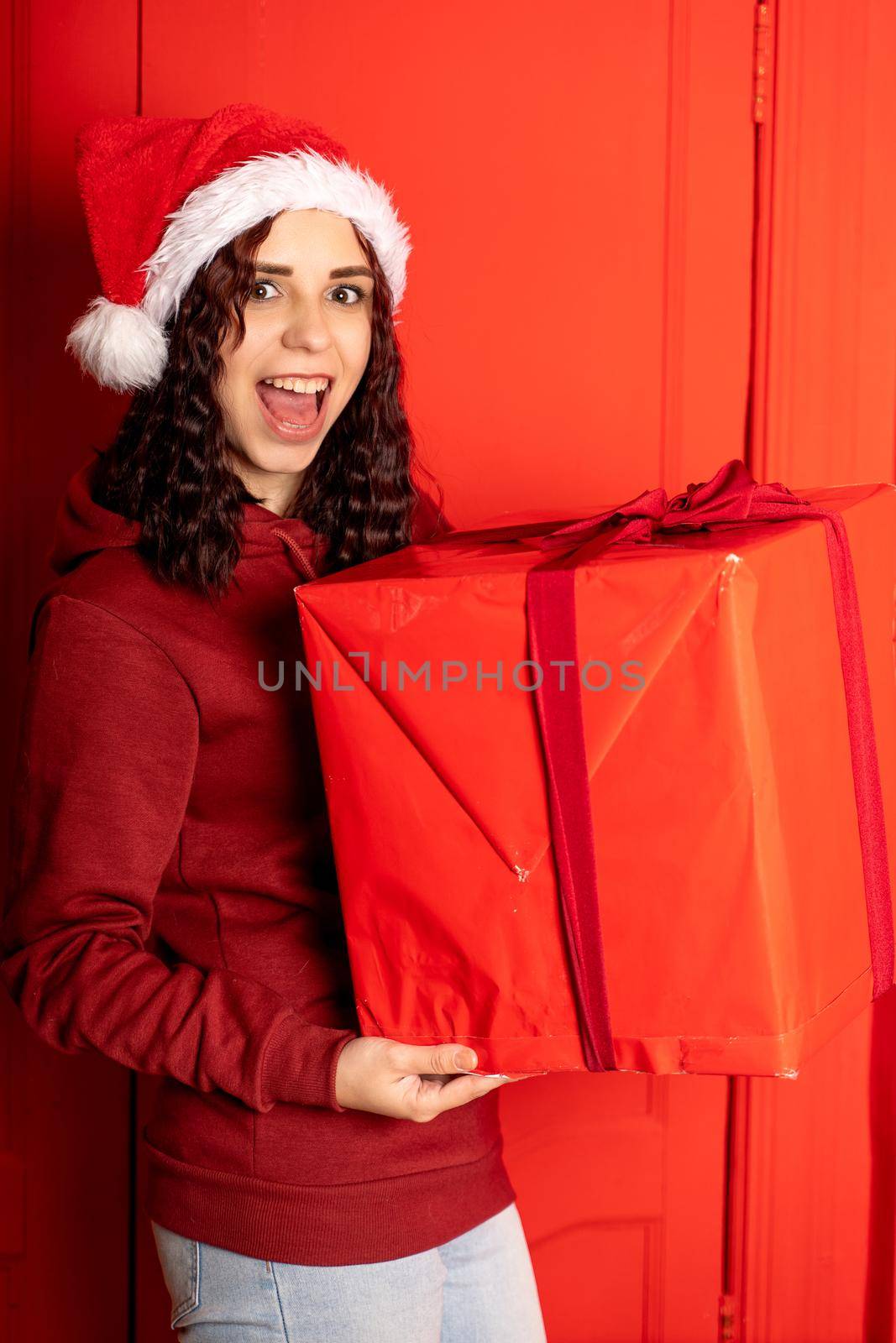 Young woman in Santa Claus hat holds big gift, standing near red wall. Happy female in Christmas hat with box of gift. Concept of holidays, presents and good mood