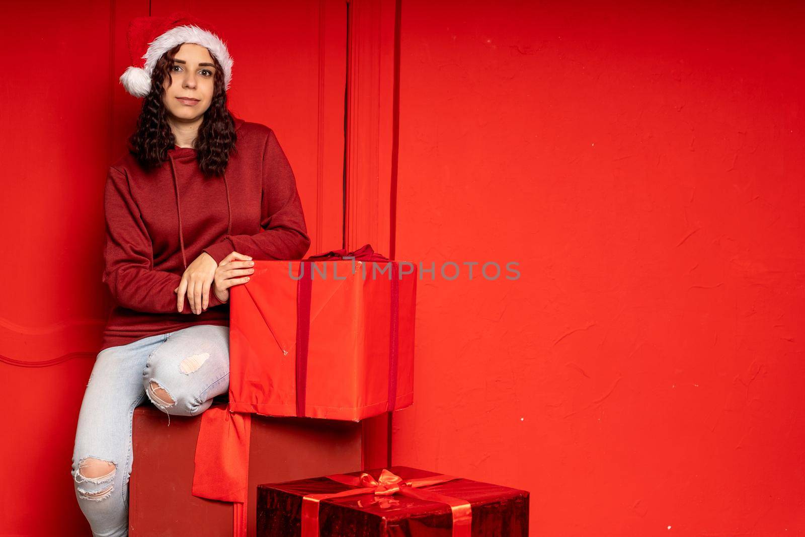 Young attractive woman in Santa Claus hat sits on gifts near red wall. Smiley female sitting on big box of gift. Concept of holidays, gifts and good mood. by epidemiks