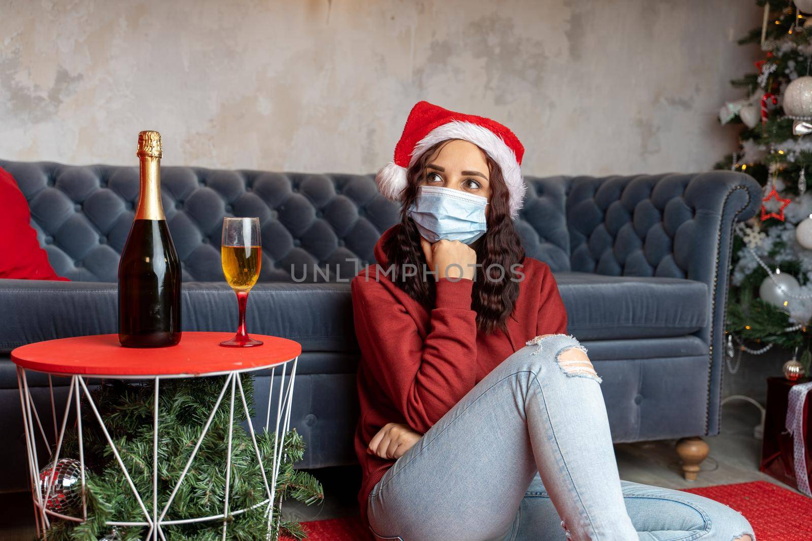 Young woman in medical mask and Santa Claus hat sitting on floor near sofa in room. Alone charming brunette in protective mask celebrating of safe Christmas during coronavirus pandemic