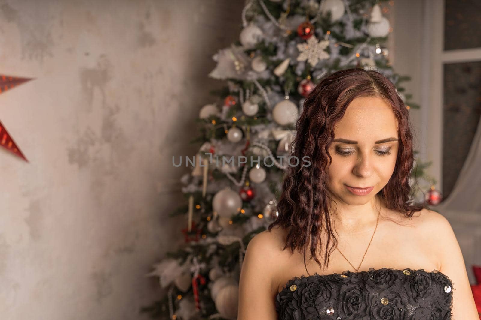 Portrait of charming brunette in black dress near Christmas tree. Young attractive woman posing at coniferous tree with decorative adornments. Concept of Christmas celebration at home