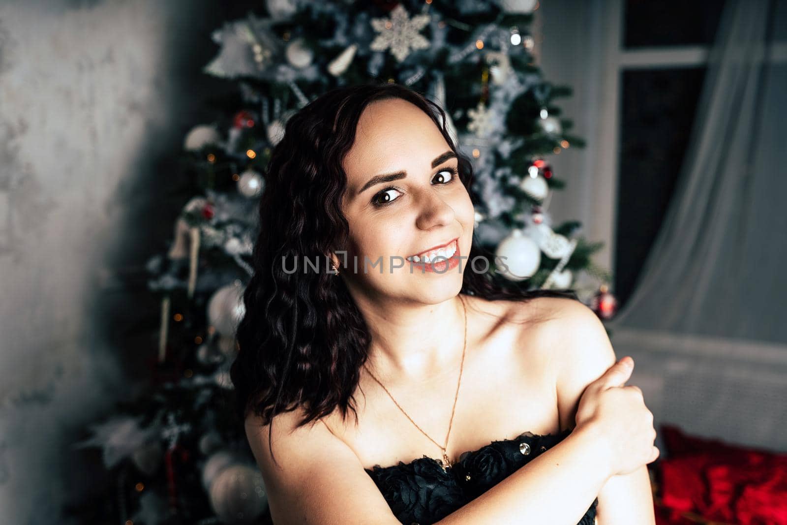 Portrait of charming brunette in black dress near Christmas tree. Young attractive woman posing at coniferous tree with decorative adornments. Concept of Christmas celebration at home