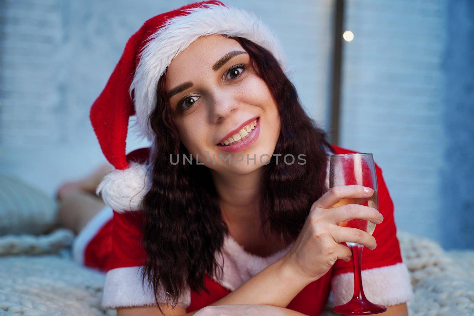 Young woman in Santa Claus costume with glass of champagne lying on bed. Charming female resting and celebrating Christmas at home. Concept of holidays and good mood