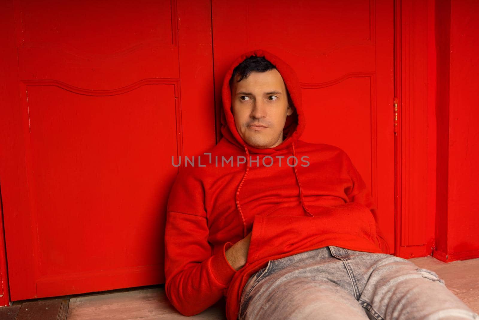 Young handsome male in hood lying on floor. Thoughtful man poses near red wall