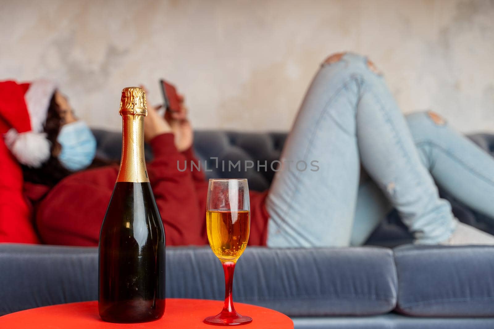 Close up of bottle and glass of champagne on table. Woman in medical mask and Santa Claus hat browses smartphone, lying on couch in background Concept of safe Christmas celebration during coronavirus