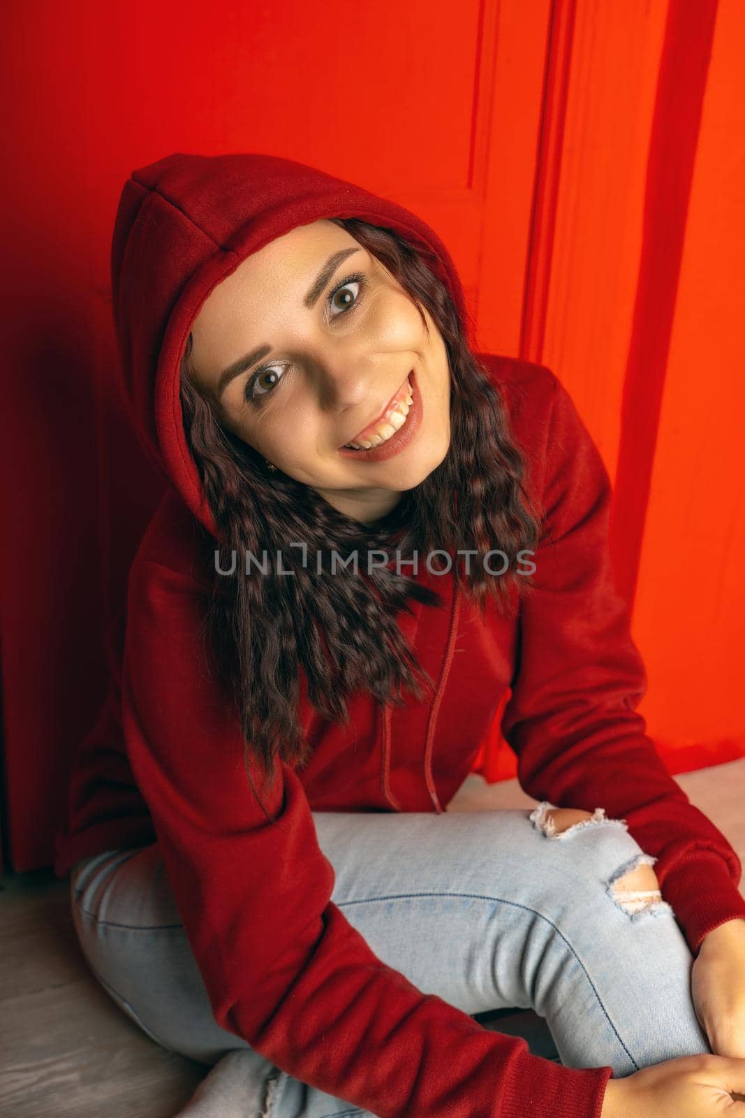 Young woman in hood sitting on floor. Curly brunette poses near red wall. by epidemiks