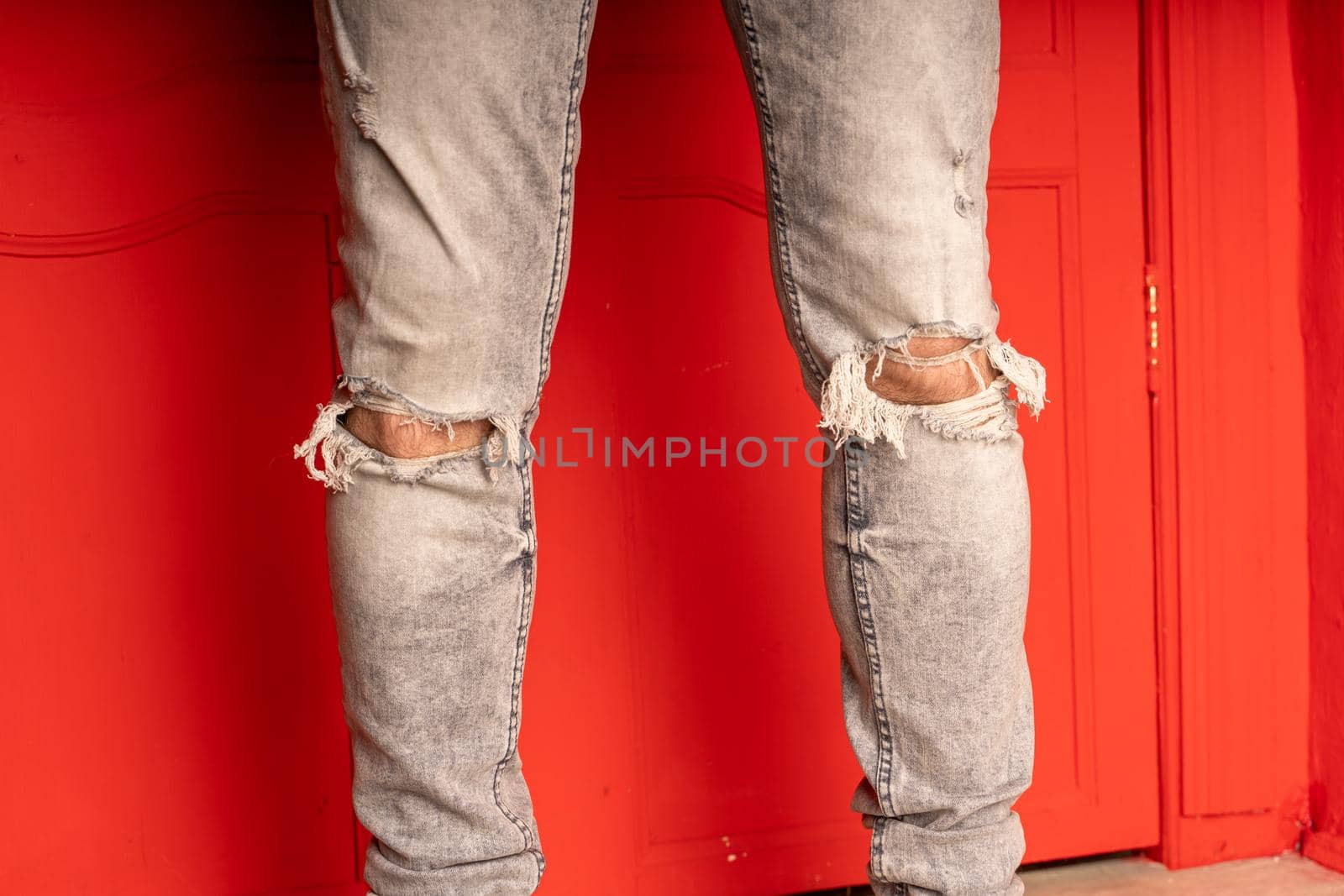 Close up of men's legs in jeans with holes on red background. Fashionable holes in grey jeans on unrecognizable man
