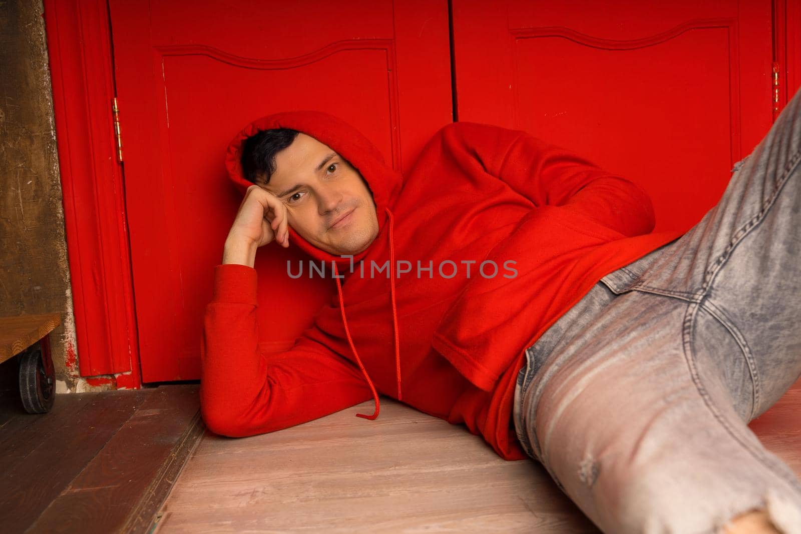 Young handsome male in hood lying on floor. Thoughtful man poses near red wall