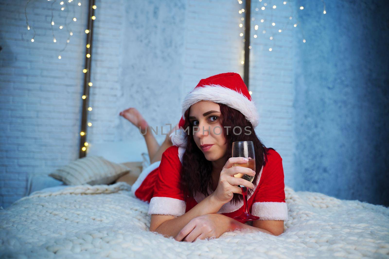 Young woman in Santa Claus costume with glass of champagne lying on bed. Charming female resting and celebrating Christmas at home. Concept of holidays and good mood