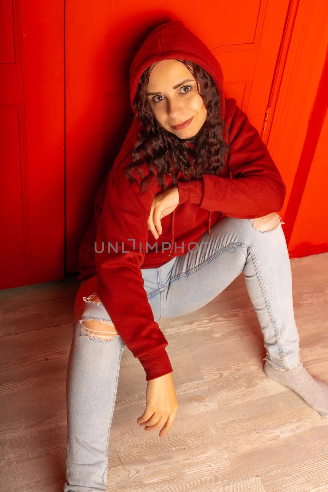 Young woman in hood sitting on floor. Curly brunette poses near red wall