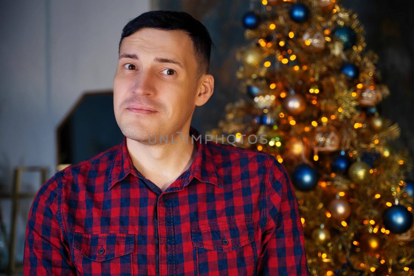 Young handsome man in plaid shirt on background of Christmas tree. Adult attractive male posing at coniferous tree with decorative adornments. Concept of Christmas celebration at home. by epidemiks