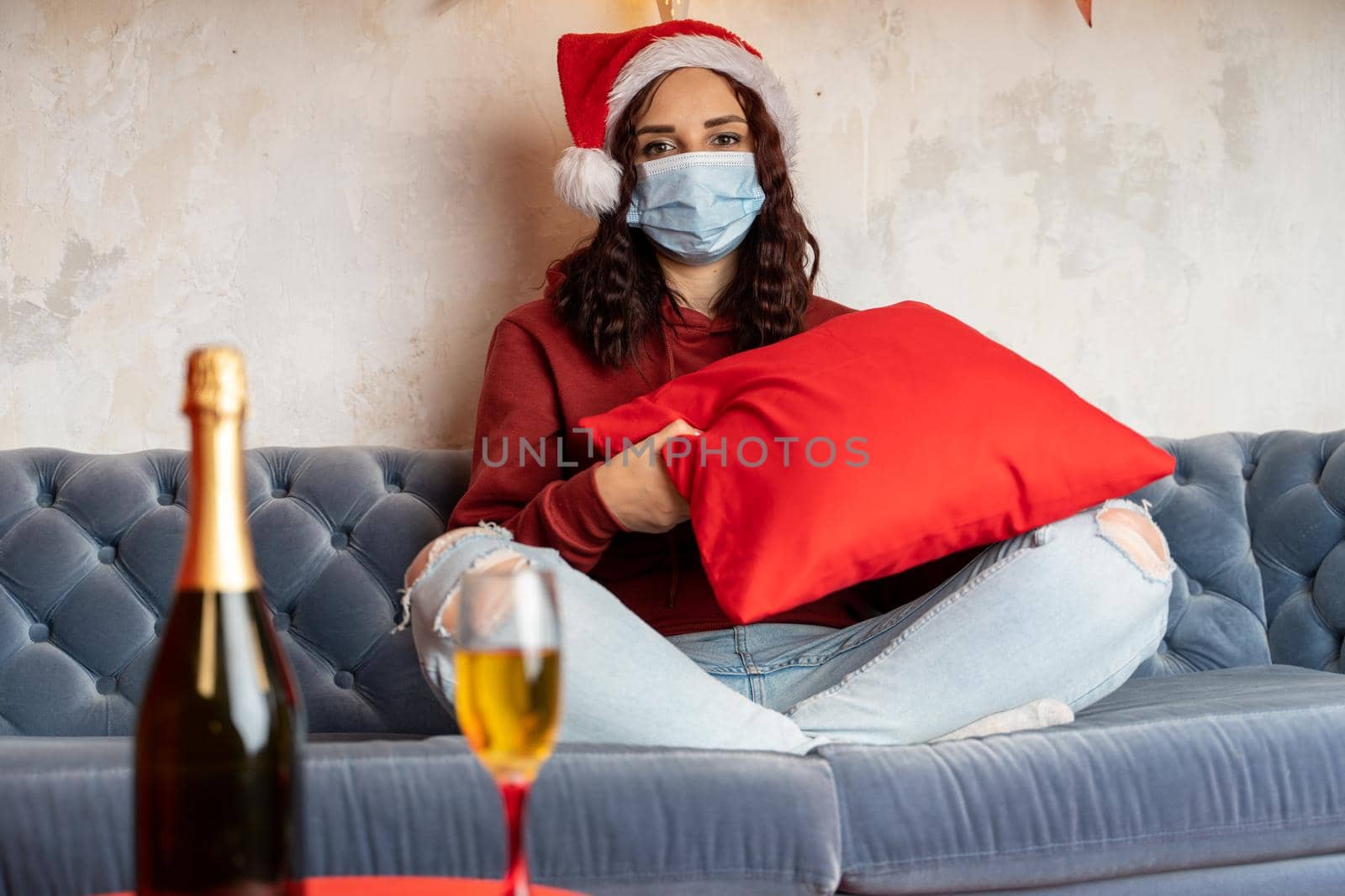 Young woman in medical mask and Santa Claus hat sitting on sofa in room. Alone charming brunette in protective mask celebrating of safe Christmas during coronavirus pandemic