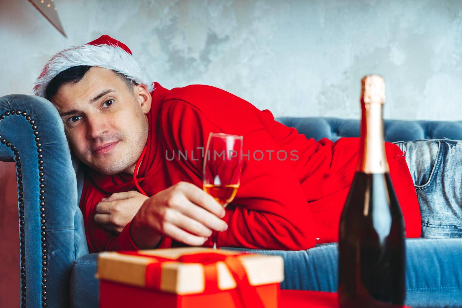 Young man in santa hat with glass of champagne lying on sofa. Adult male resting in room. Concept of Christmas celebration at home. by epidemiks