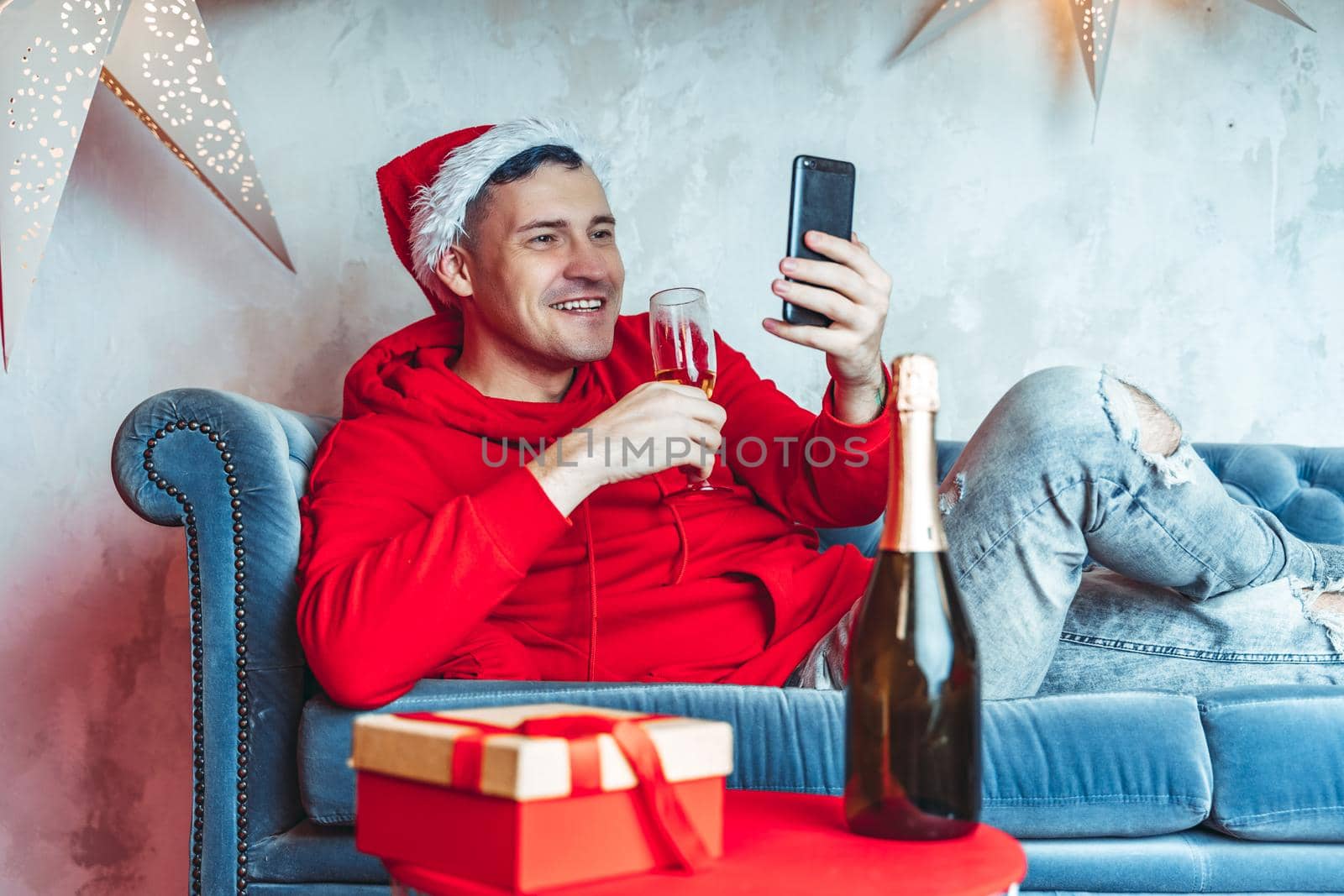 Young handsome man in Santa Claus hat with glass of champagne conducting live broadcast with friends on smartphone. Happy guy celebrating Christmas online on internet and lying on couch at home. by epidemiks