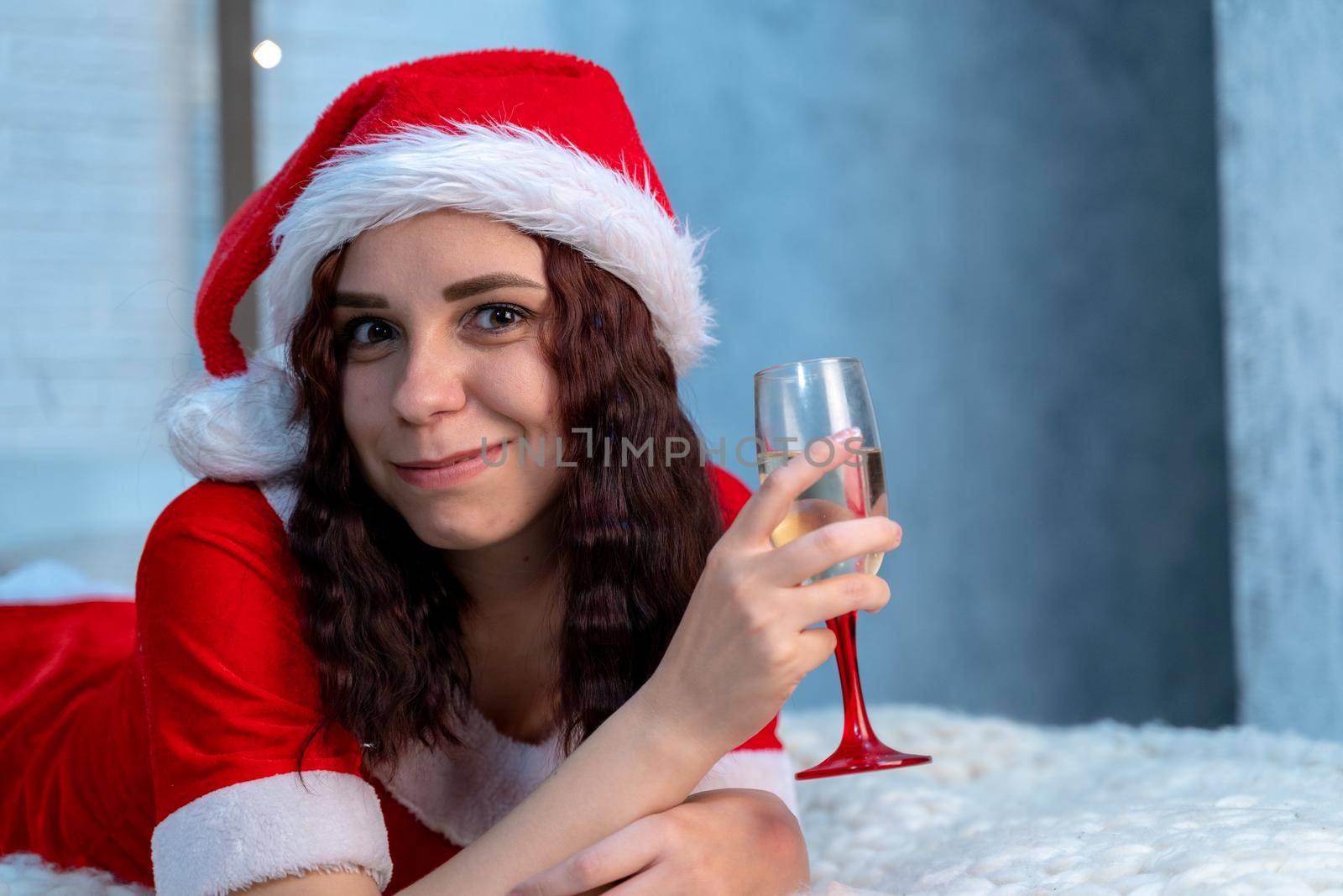 Young woman in Santa Claus costume with glass of champagne lying on bed. Charming female resting and celebrating Christmas at home. Concept of holidays and good mood