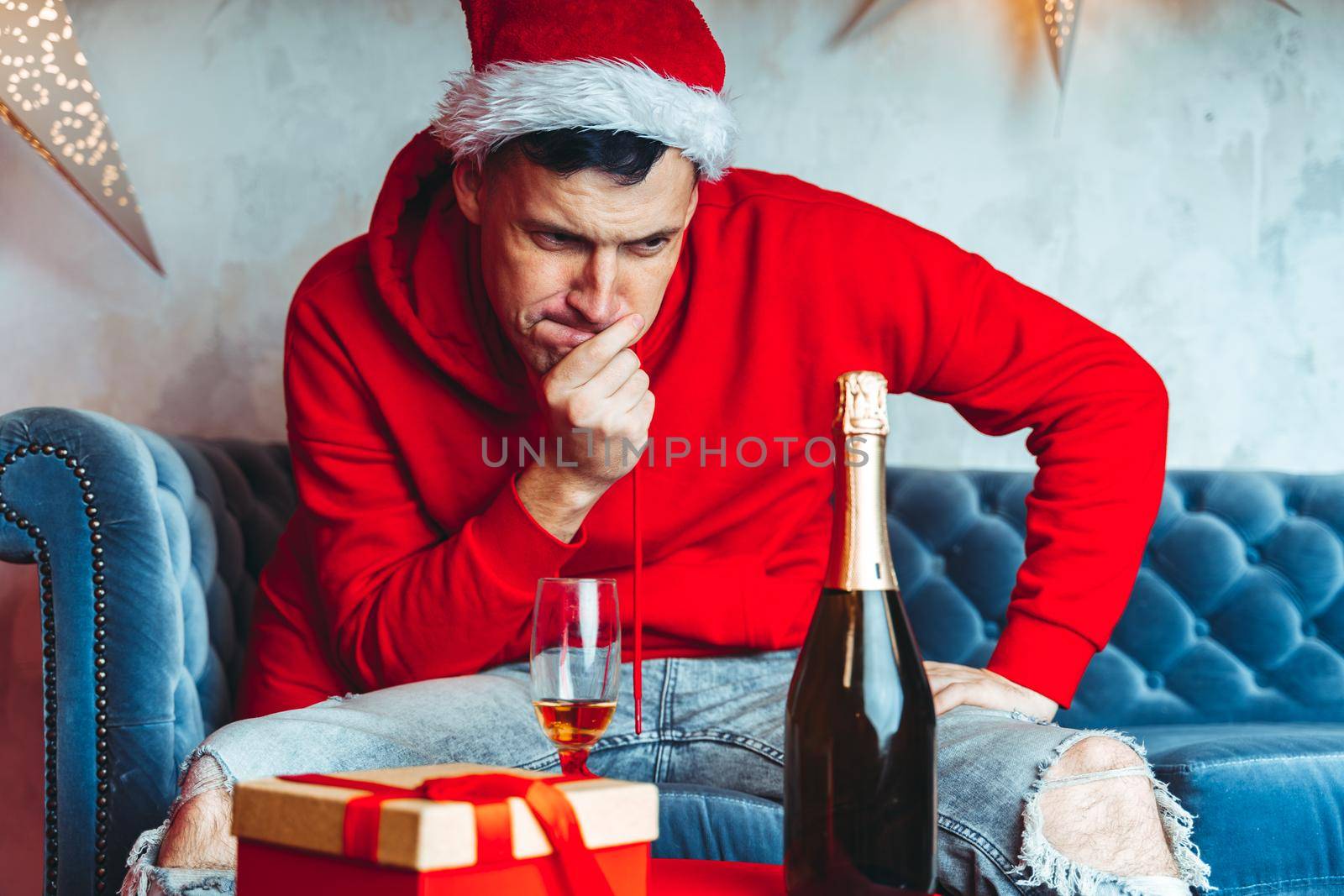 Young man in Santa hat celebrating Christmas in solitude at home. Adult guy rests, sitting on sofa in room. Concept of holidays and good mood. by epidemiks