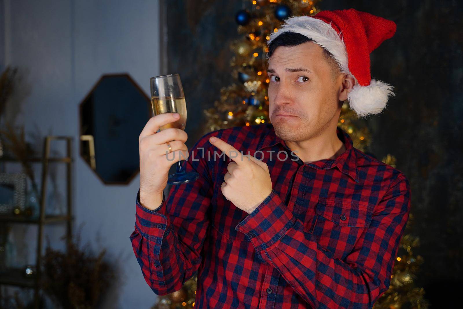Young man in santa hat with glass of champagne on background of Christmas tree. Adult male posing at coniferous tree with decorative adornments. Concept of Christmas celebration at home