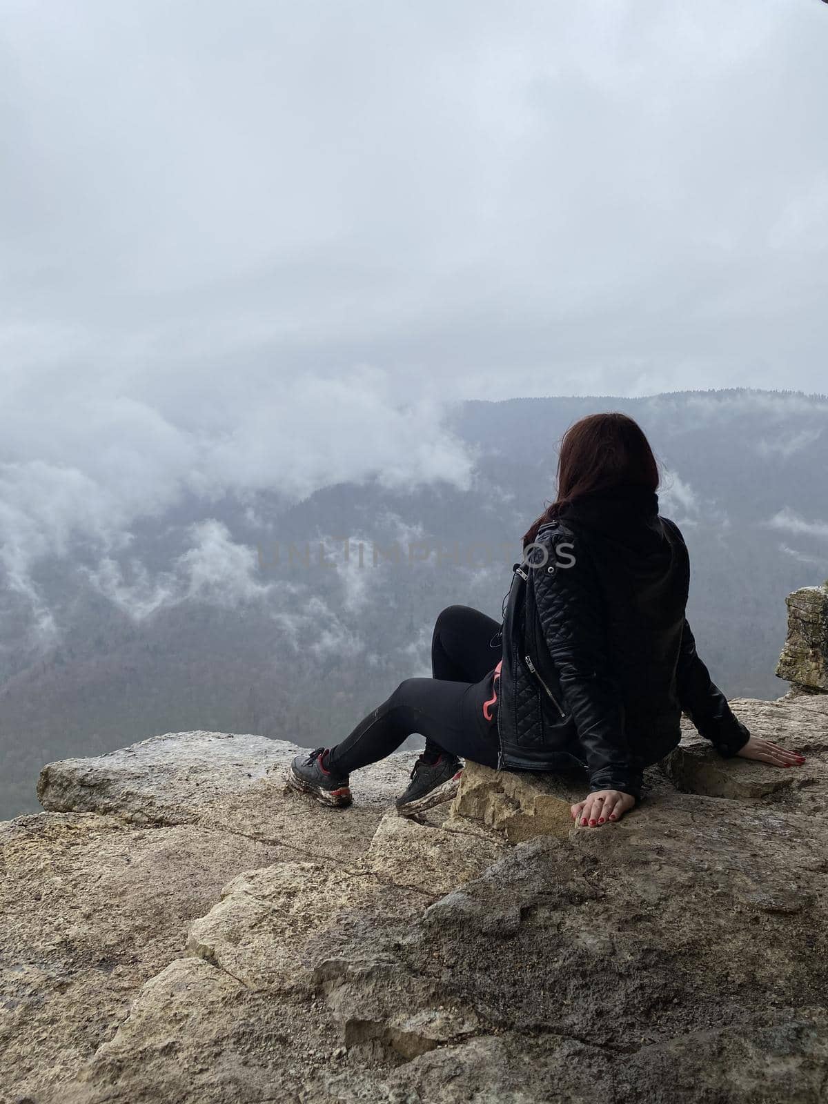 Young woman sitting on edge of cliff in foggy and cloudy weather. Female tourist enjoys spectacular view of nature, sitting on high rock