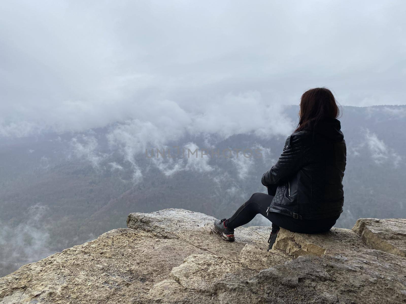 Young woman sitting on edge of cliff in foggy and cloudy weather. Female tourist enjoys spectacular view of nature, sitting on high rock. by epidemiks