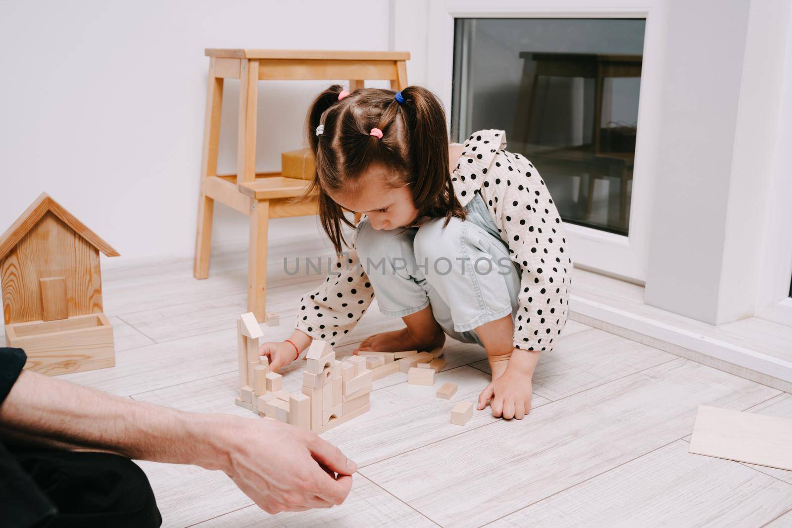 Girl sits on the floor in a room and plays with her dad in a natral wood constructor. Wooden childrens construction kit. Toys made of eco-friendly material. The castle is made of wooden cubes.