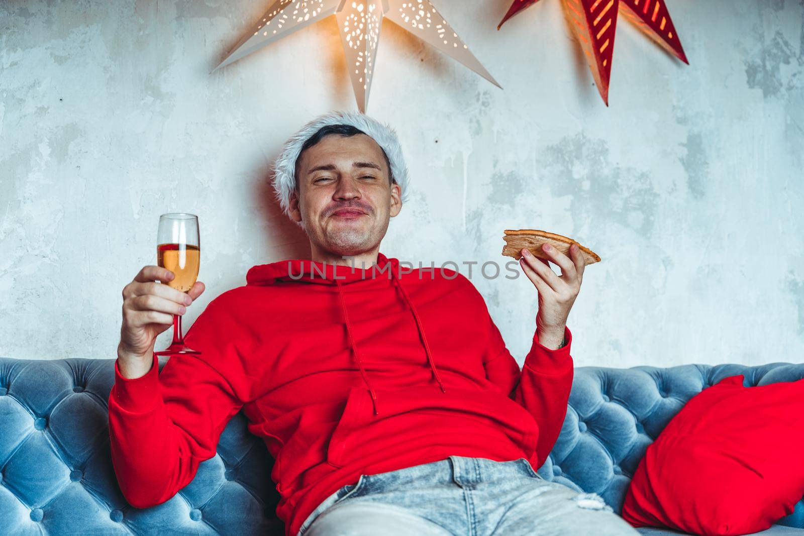 Young man in santa hat drinks champagne and eats pizza, sitting on sofa in room. Happy male resting with food and alcohol. Concept of Christmas celebration at home