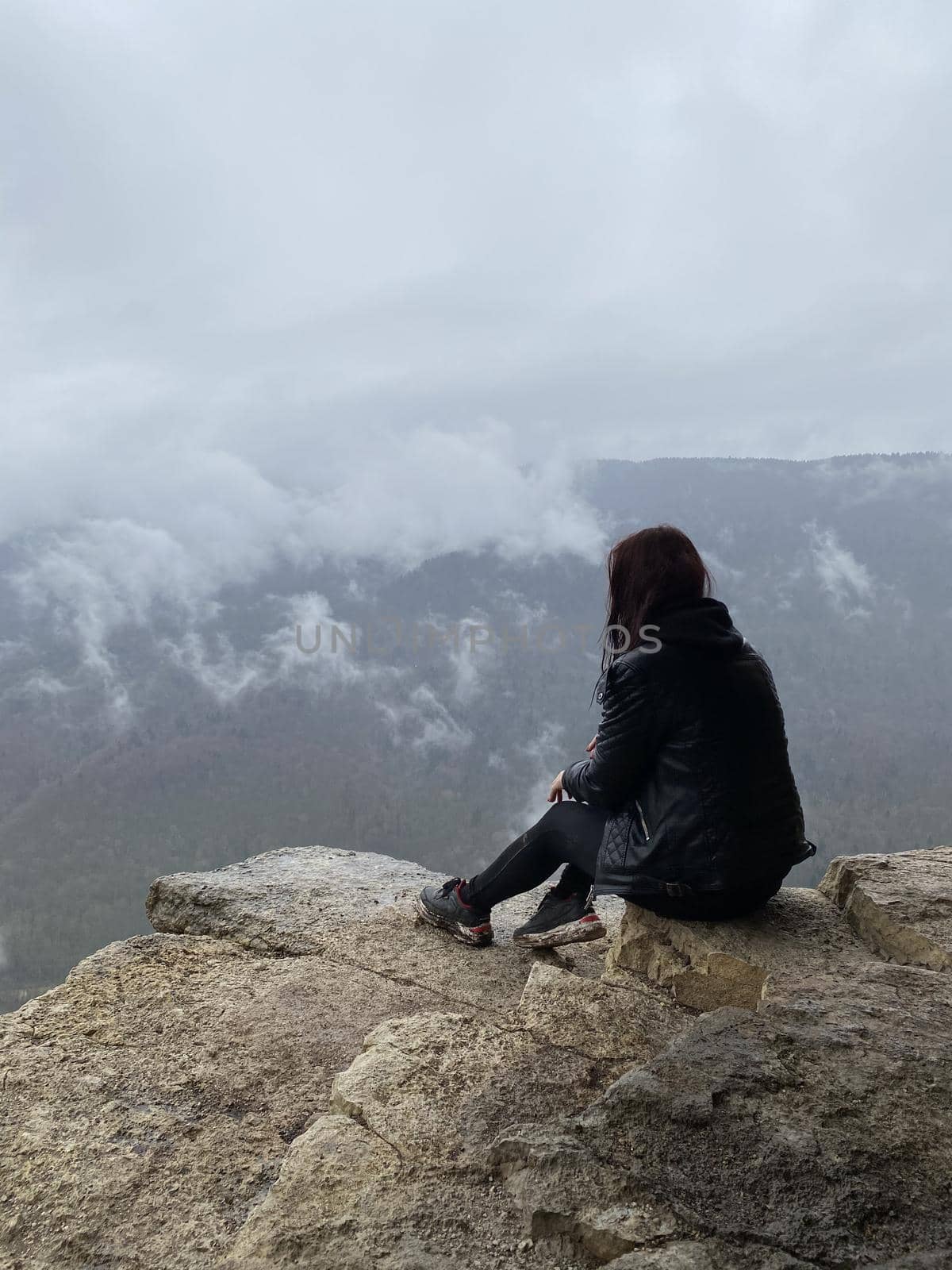 Young woman sitting on edge of cliff in foggy and cloudy weather. Female tourist enjoys spectacular view of nature, sitting on high rock