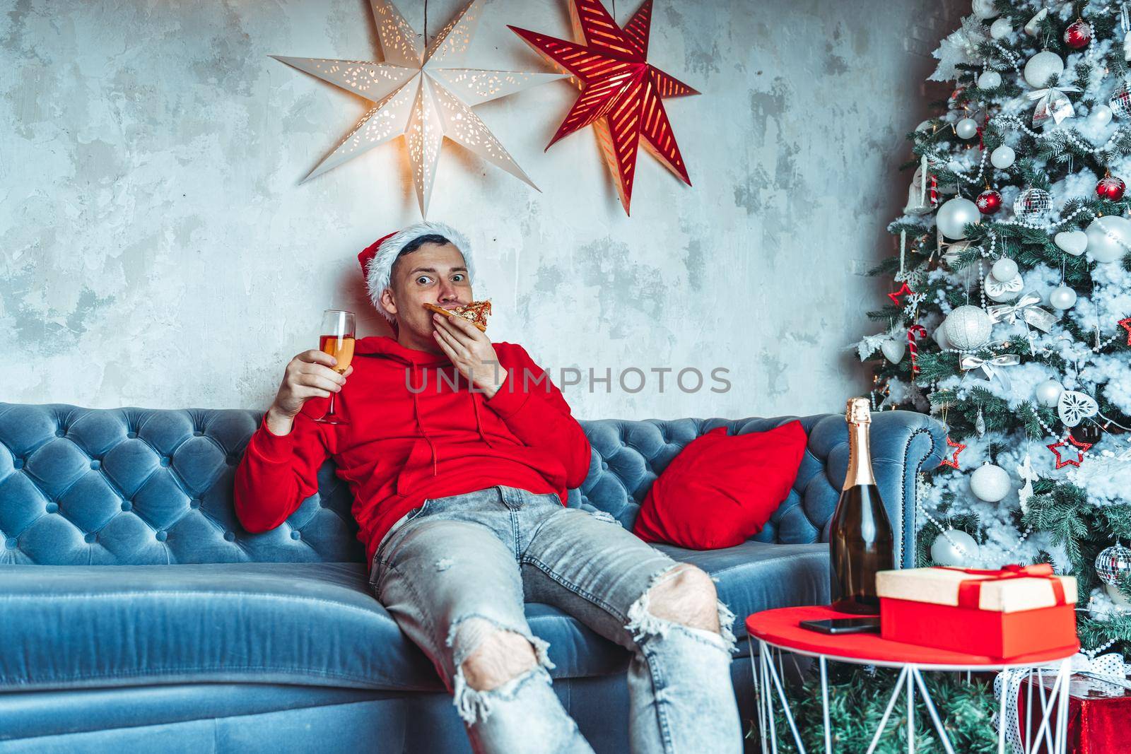 Young man in santa hat drinks champagne and eats pizza, sitting on sofa in room. Happy male resting with food and alcohol. Concept of Christmas celebration at home. by epidemiks