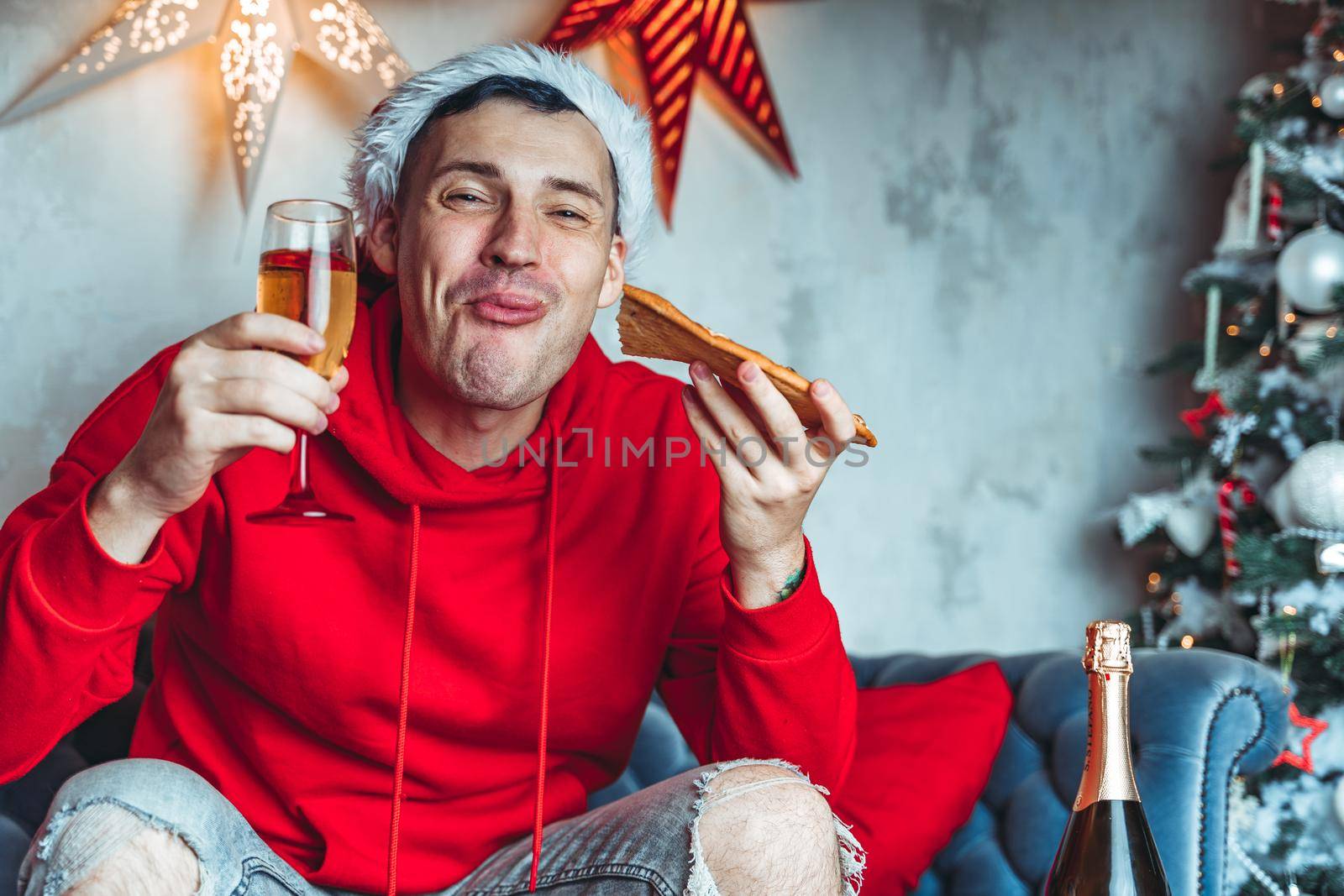 Young man in santa hat drinks champagne and eats pizza, sitting on sofa in room. Happy male resting with food and alcohol. Concept of Christmas celebration at home