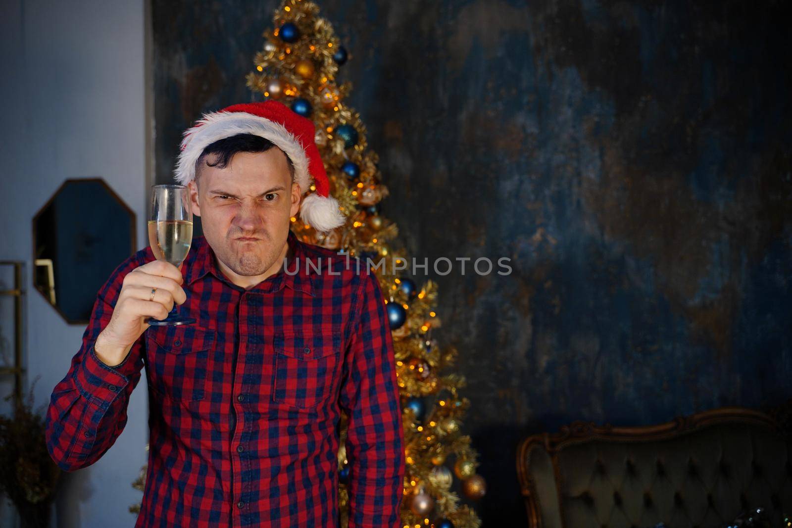 Crazy male in santa hat with glass of champagne writhes face on background of Christmas tree. Aggressive man grimaces, looking at camera in dark room