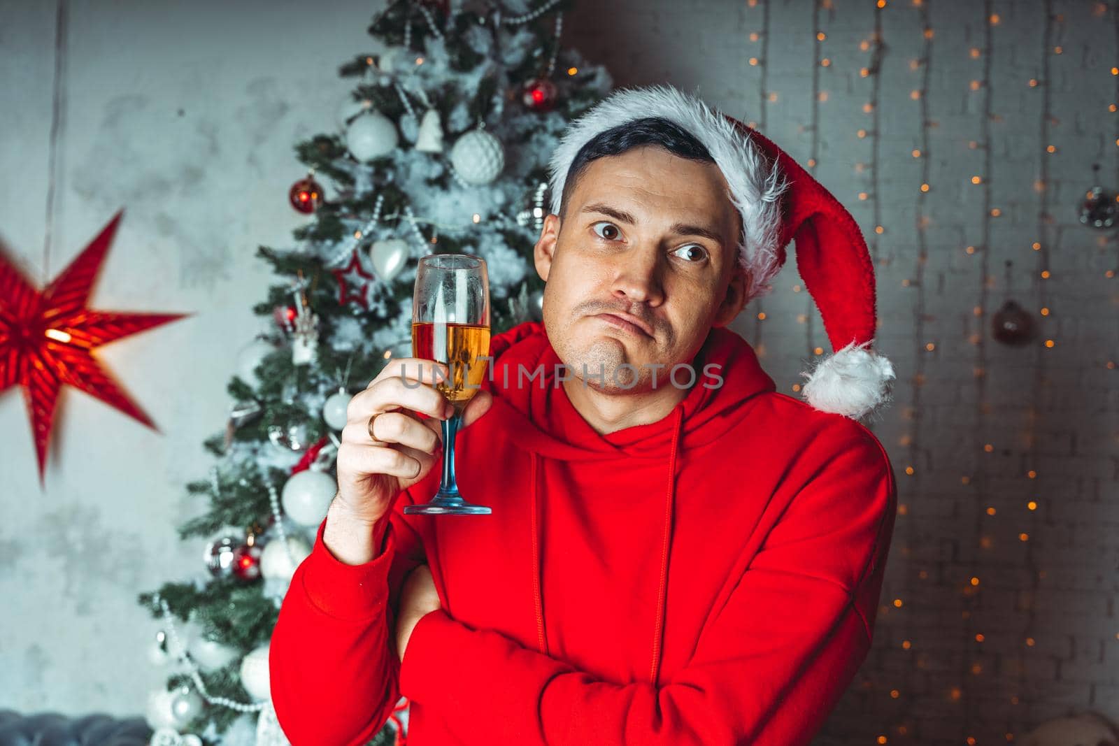 Young man in santa hat with glass of champagne on background of Christmas tree. Adult male posing at coniferous tree with decorative adornments. Concept of Christmas celebration at home. by epidemiks