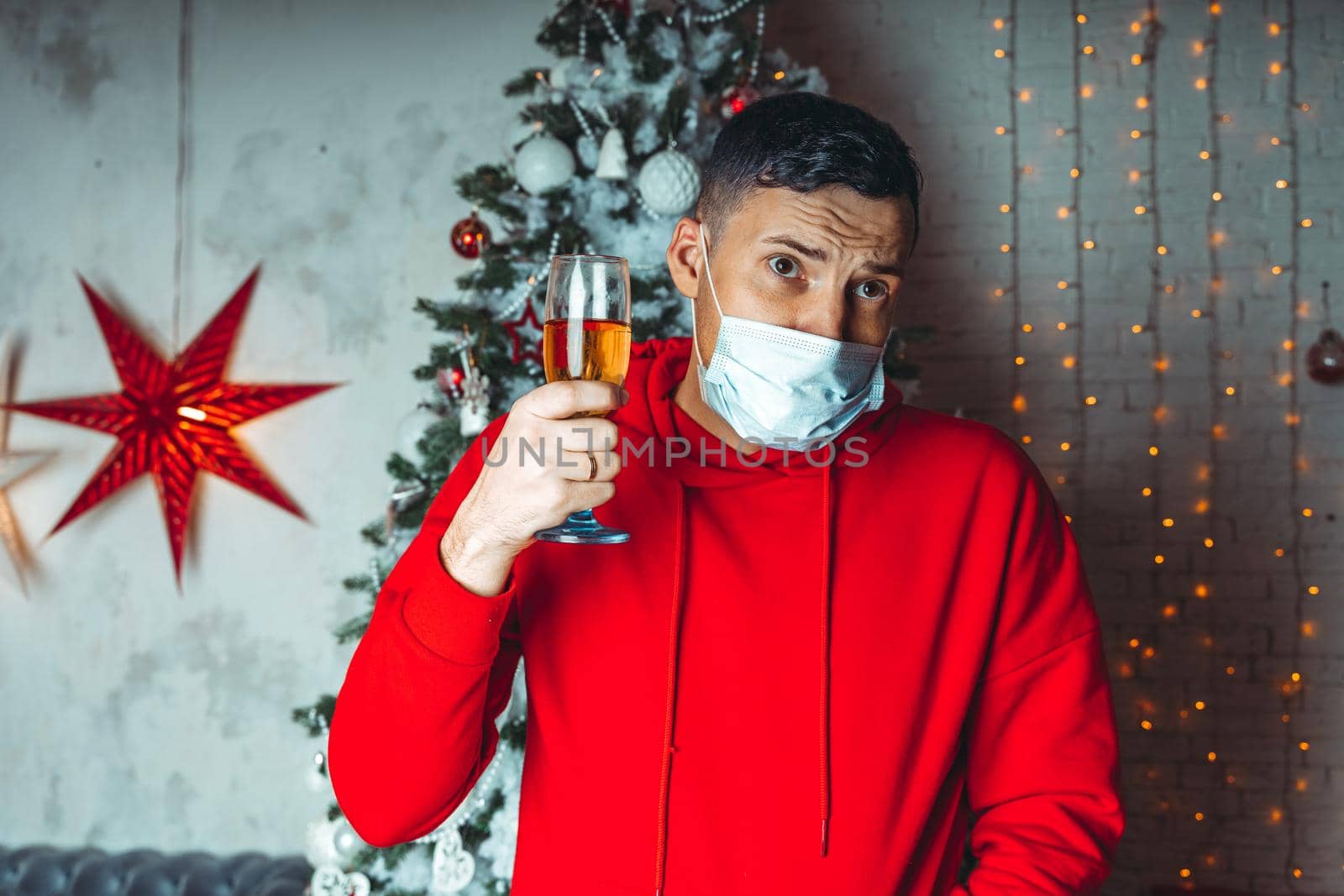 Young man in medical mask with glass of champagne on background of Christmas tree. Adult male celebrating of safe Christmas during coronavirus pandemic