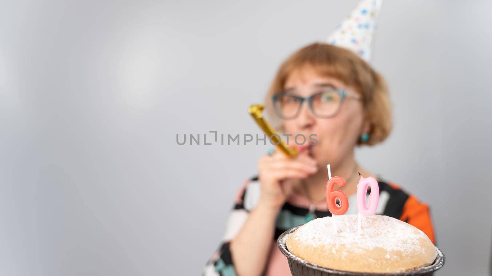 A portrait of a smiling elderly woman in a festive cap holding a cake with candles in the form of the number 60. Anniversary.