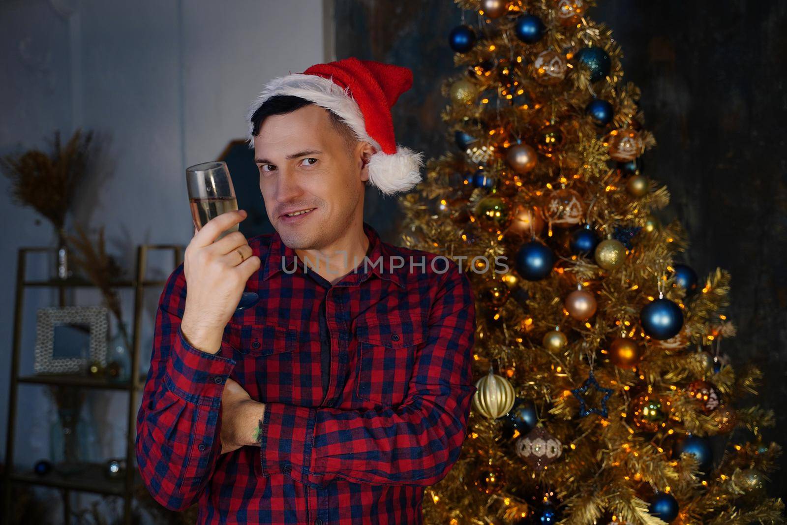 Young man in santa hat with glass of champagne on background of Christmas tree. Adult male posing at coniferous tree with decorative adornments. Concept of Christmas celebration at home by epidemiks