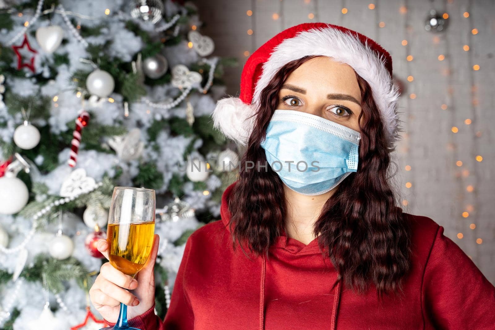 Young woman in medical mask and santa hat with glass of champagne on background of Christmas tree. Charming brunette in protective mask celebrating of safe Christmas during coronavirus pandemic. by epidemiks