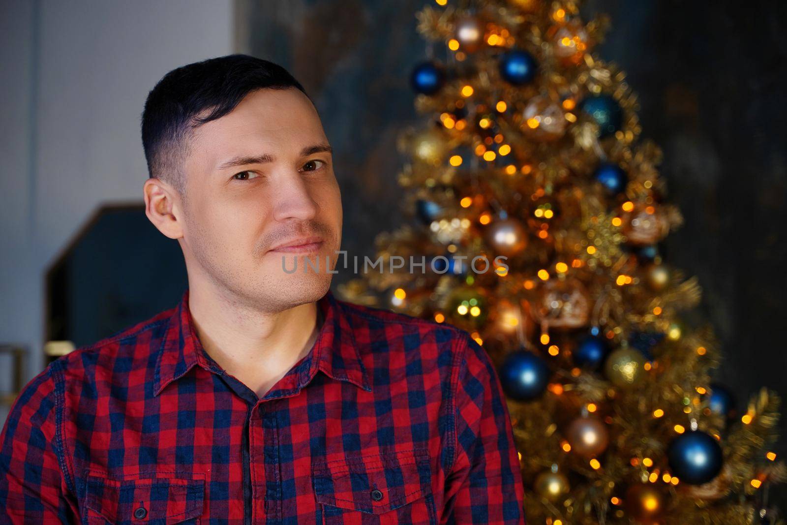 Young handsome man in plaid shirt on background of Christmas tree. Adult attractive male posing at coniferous tree with decorative adornments. Concept of Christmas celebration at home