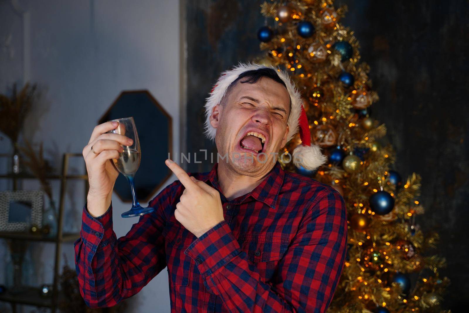 Young man in santa hat drinks champagne and grimaces from alcohol on background of Christmas tree in dark room. Concept of Christmas celebration at home