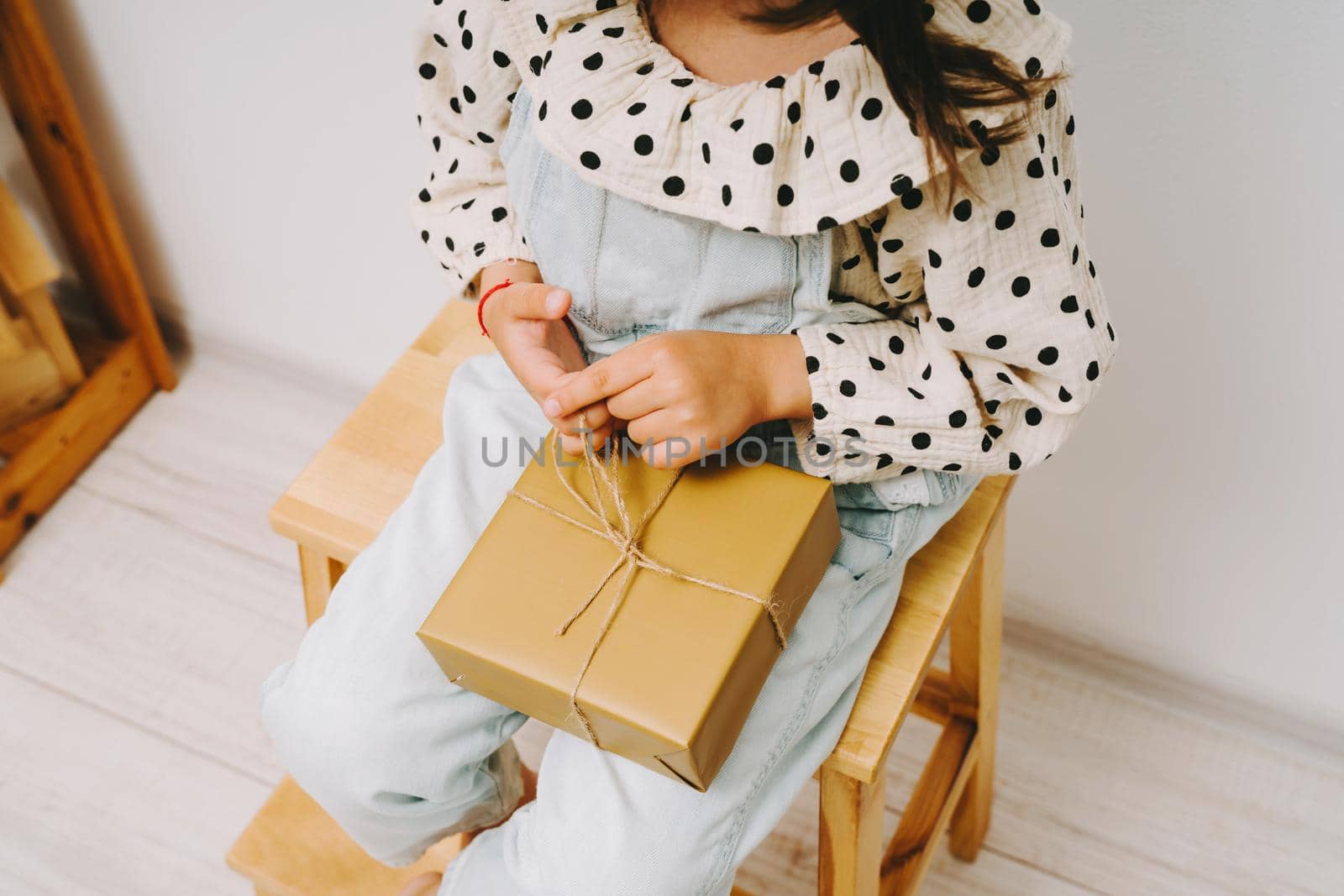 A little girl in a denim jumpsuit sits on a wooden chair and holds a gift box on her knees.