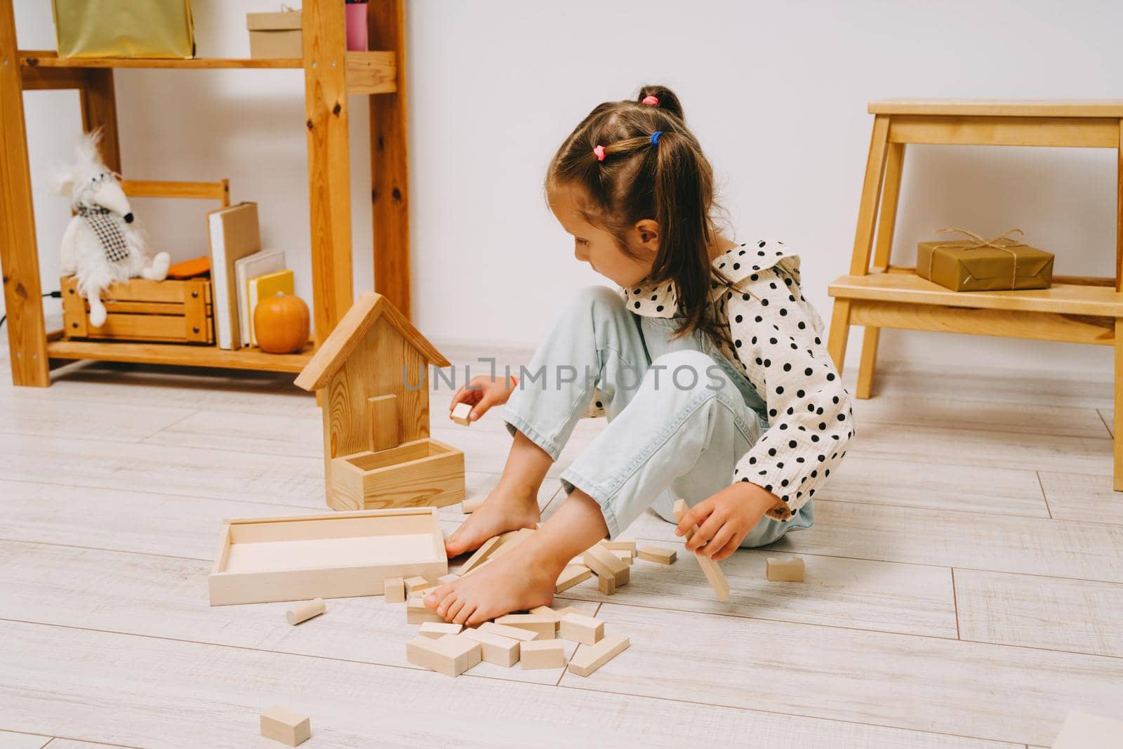 A girl with two ponytails on her head sits on the floor in her room and plays with wooden cubes. The girl sits on the floor and builds a tower out of the constructor.