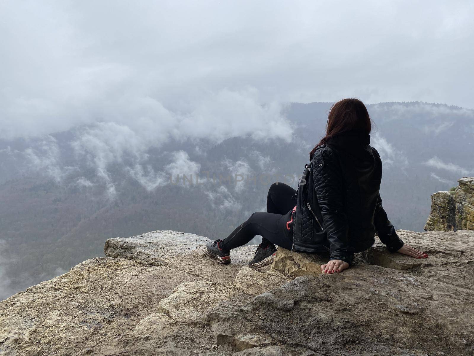 Young woman sitting on edge of cliff in foggy and cloudy weather. Female tourist enjoys spectacular view of nature, sitting on high rock. by epidemiks