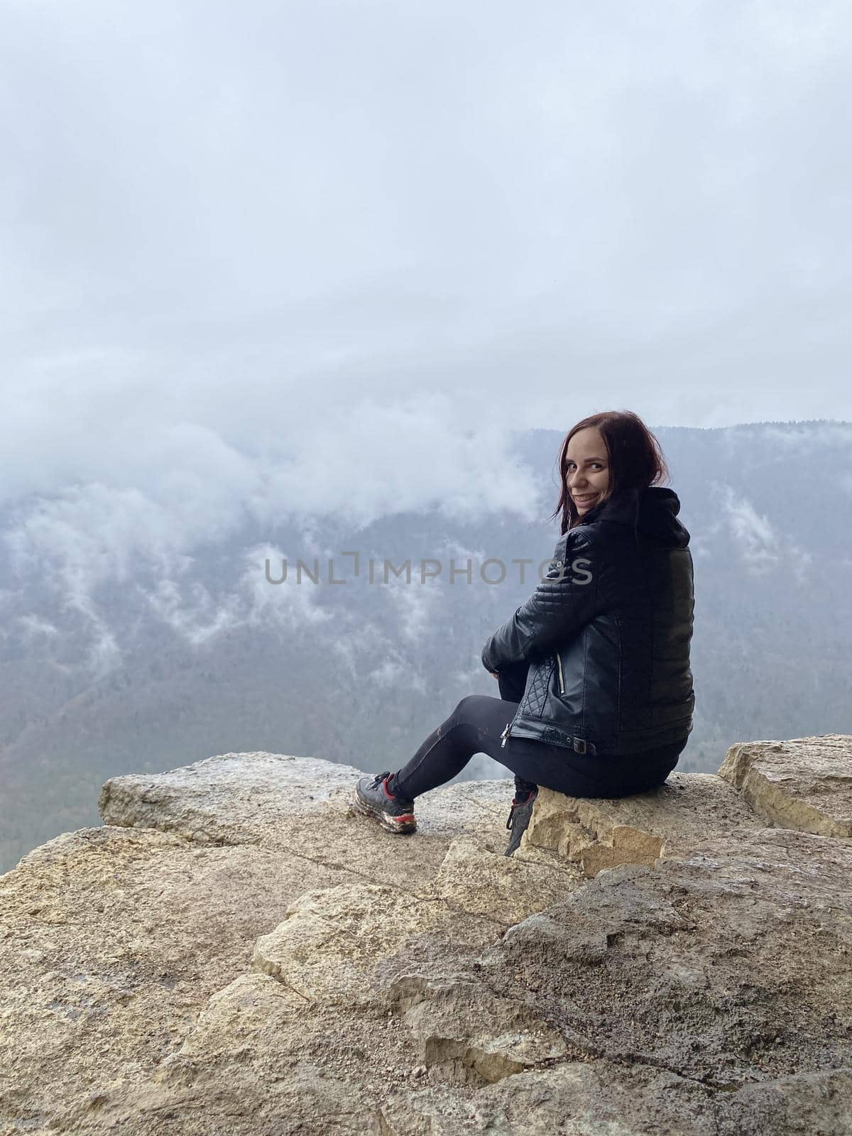Young woman sitting on edge of cliff in foggy and cloudy weather. Female tourist enjoys spectacular view of nature, sitting on high rock. by epidemiks