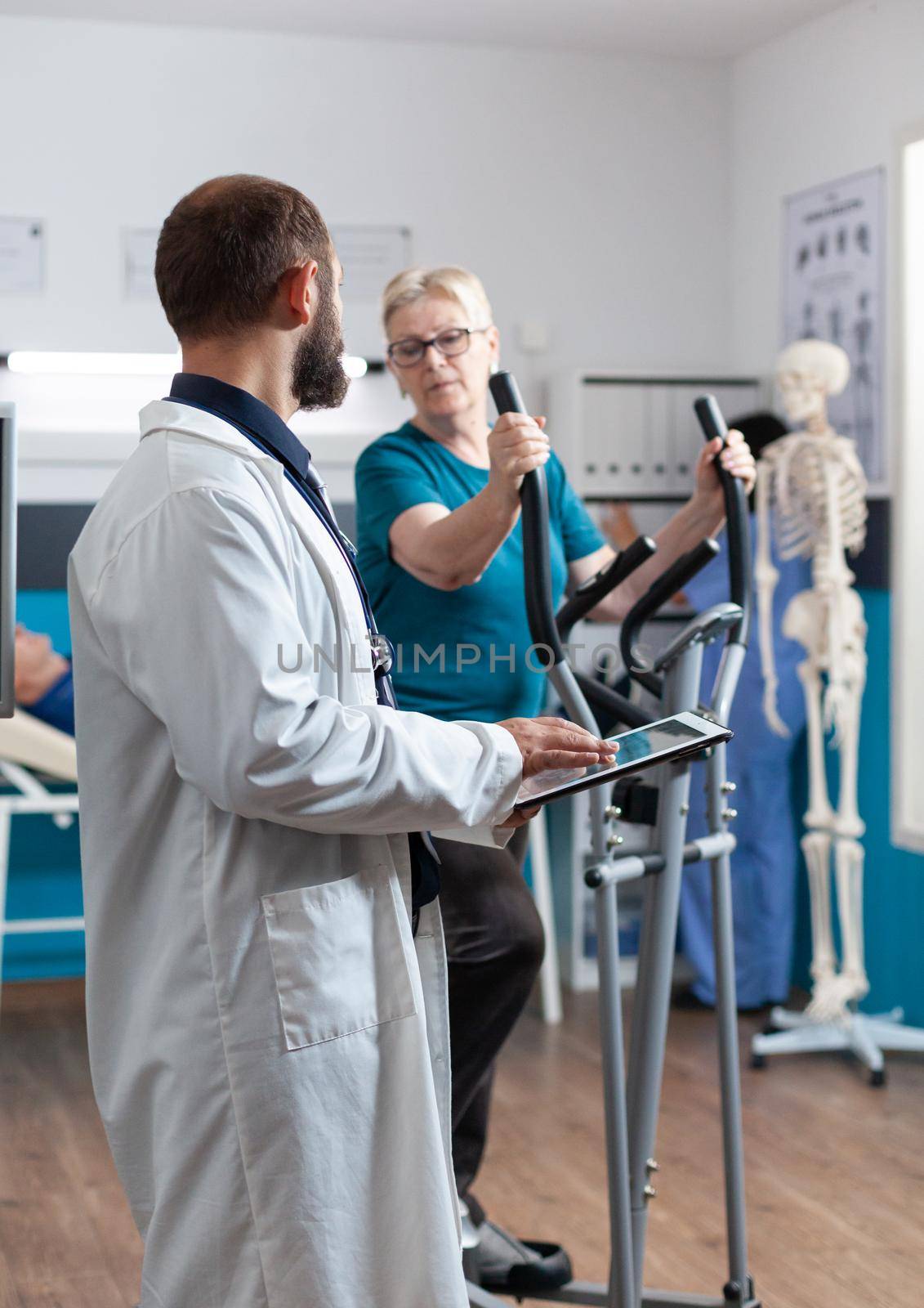 Doctor holding digital tablet for checkup with woman at recovery. Senior patient using electrical bicycle for physical exercise and activity while talking to medic about physiotherapy