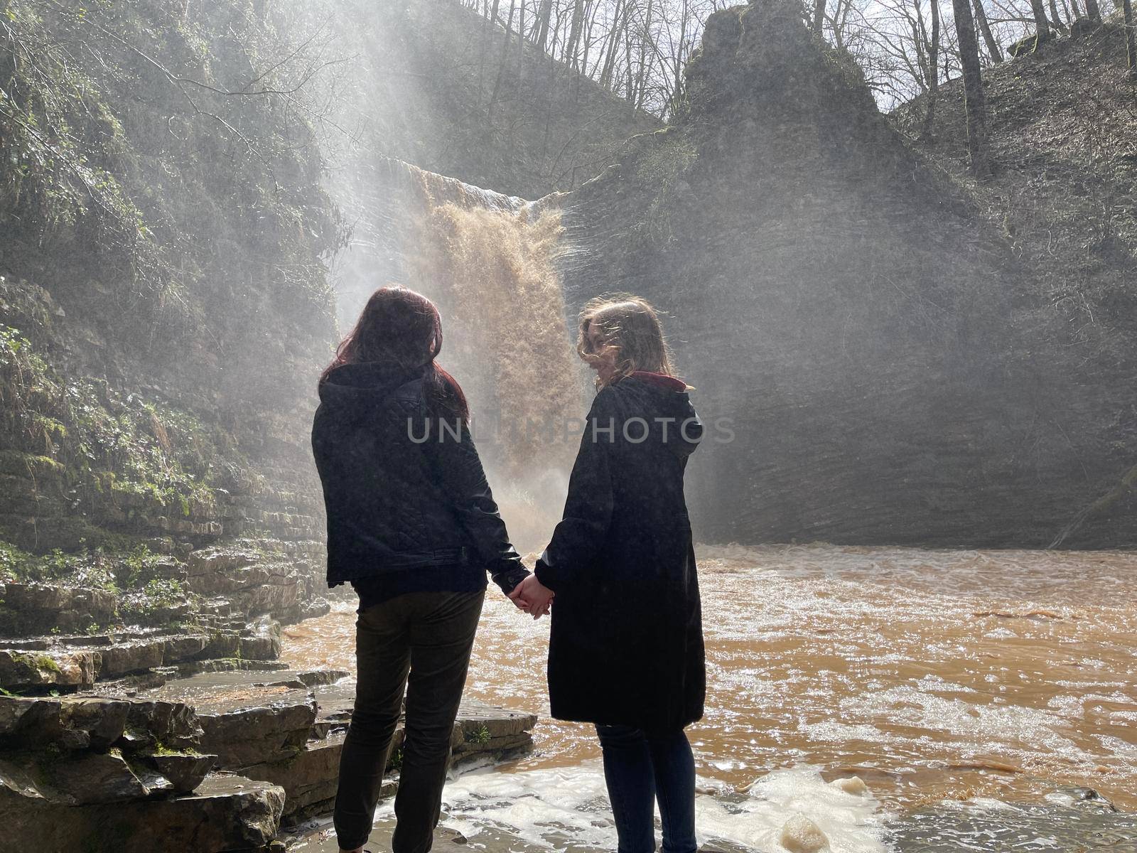 Rear view of two women looking on powerful muddy waterfall. Tourists standing on rocks in mountainous terrain and enjoying beautiful view of cataract