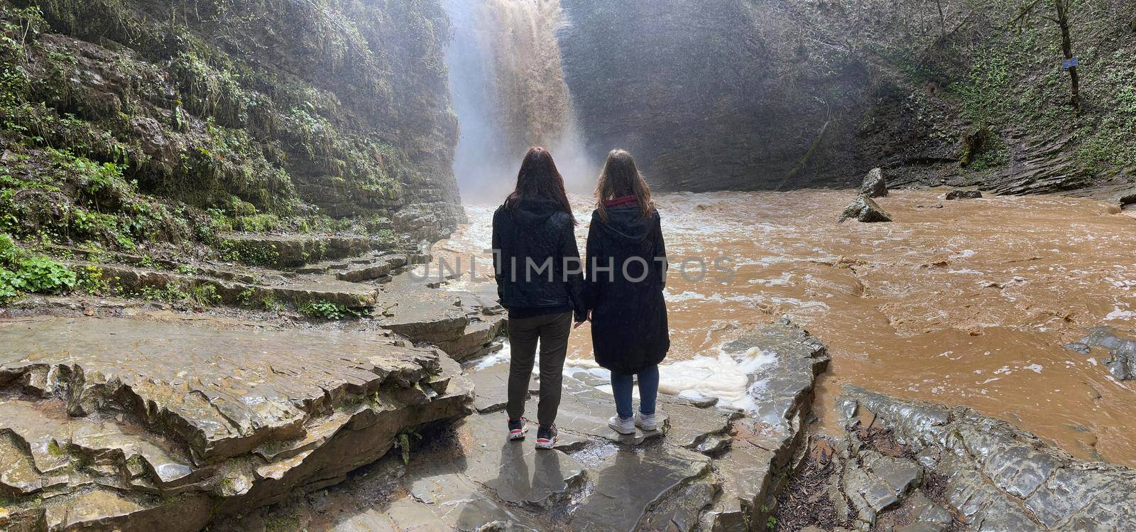 Rear view of two women looking on powerful muddy waterfall. Tourists standing on rocks in mountainous terrain and enjoying beautiful view of cataract. by epidemiks