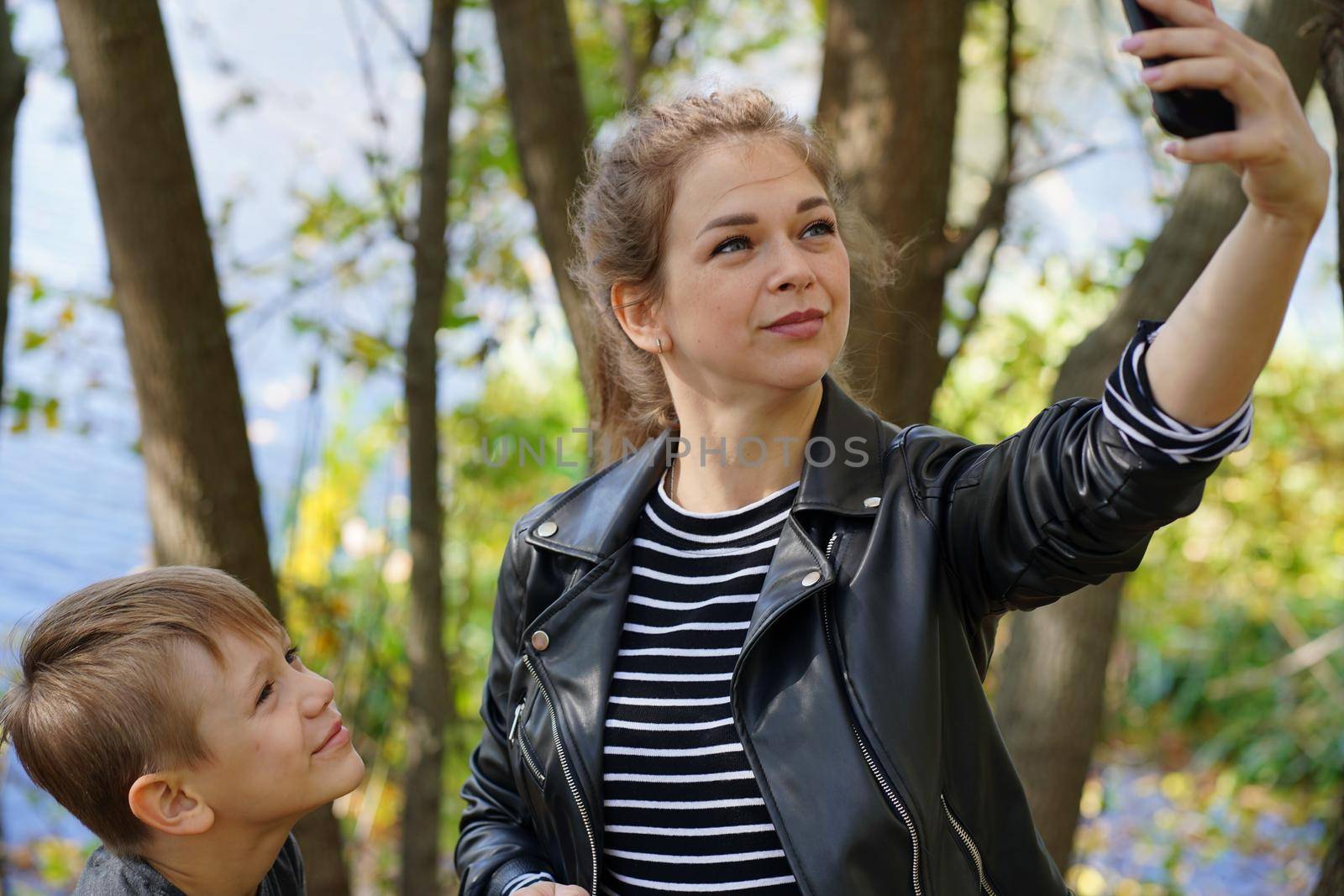 A young mother with her son takes a selfie. Smiling young woman with long hair and a child take a selfie with a smartphone, in the park.