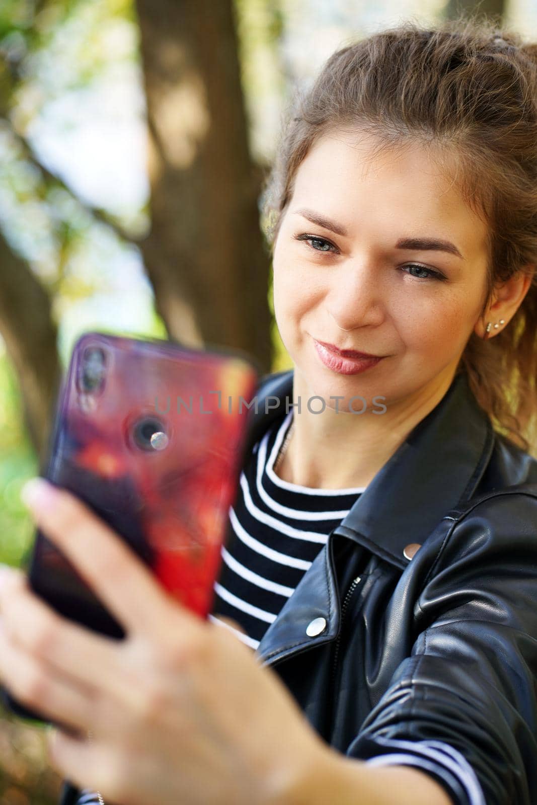 Young woman using smartphone in park. Smiling young female with long hair waving in wind browsing mobile phone while spending autumn day in park.