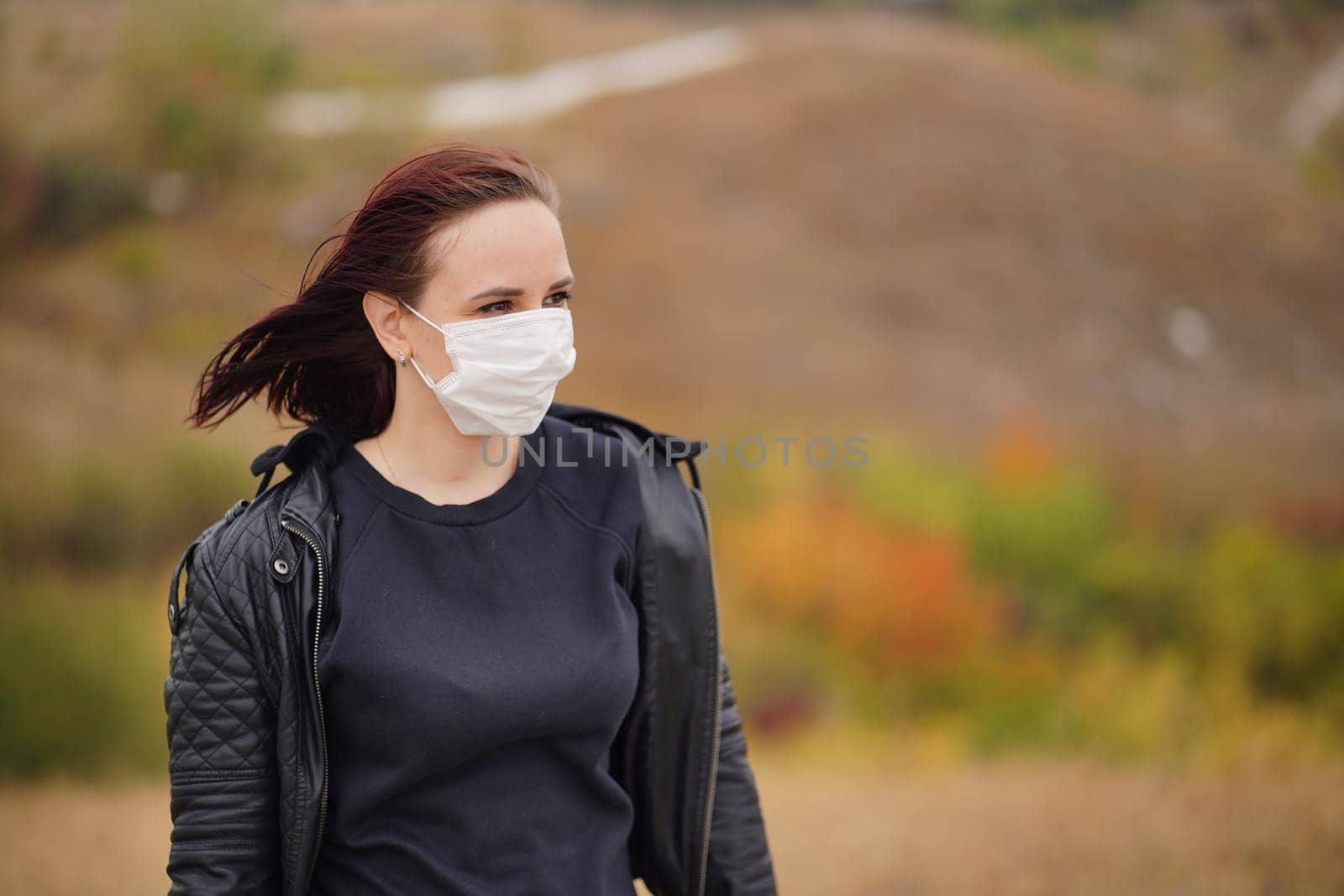 Young woman in medical mask standing in countryside. Adult brunette in protective mask in cool weather on walk in hilly terrain. by epidemiks