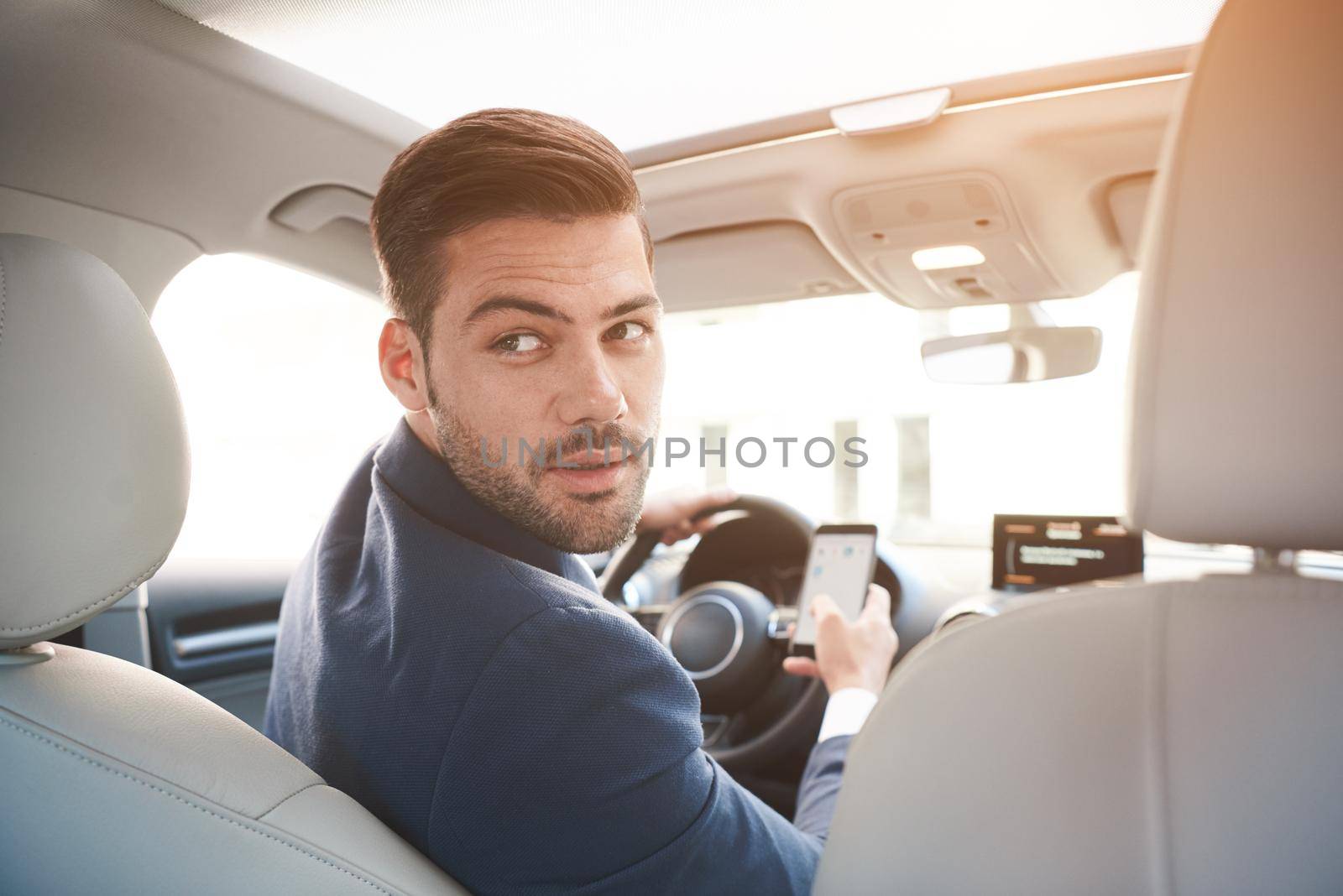 Portrait of a stylish confident businessman in a car wearing suit. Turning to the rear seats with a serious expression and phone in hand, park the car.