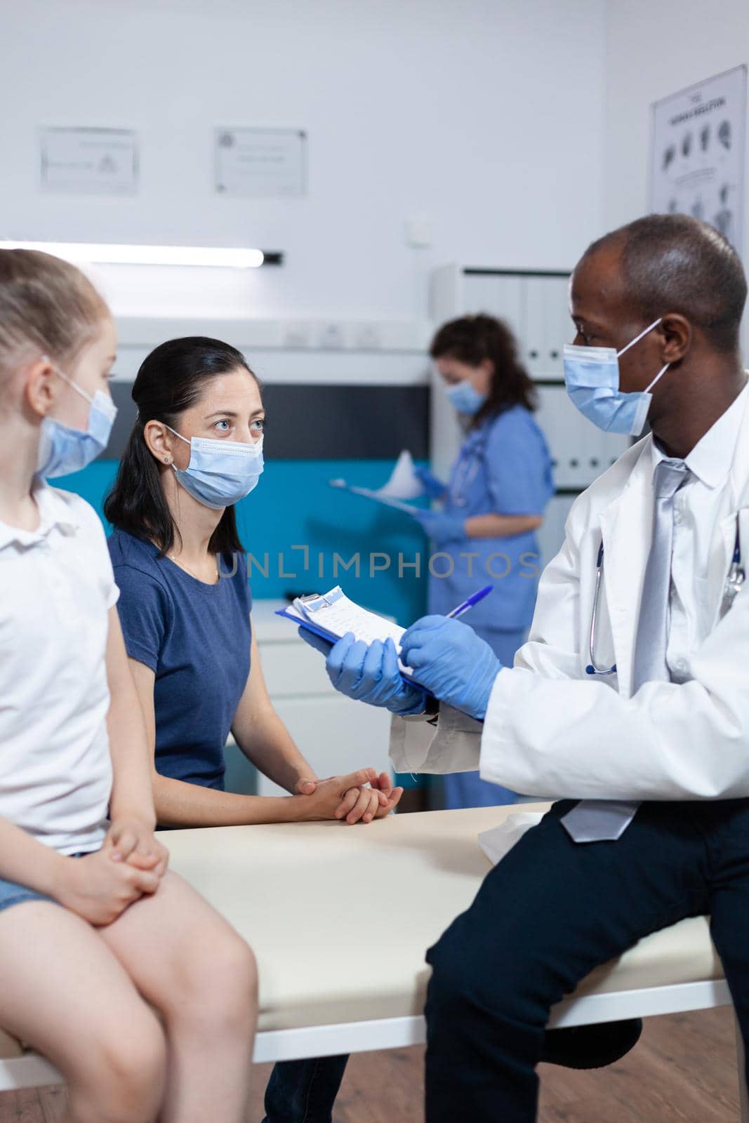 African american pediatrician doctor writing disease symptoms on clipboard discussing healthcare treatment with family during clinical examination in hospital office. Medical team with face mask
