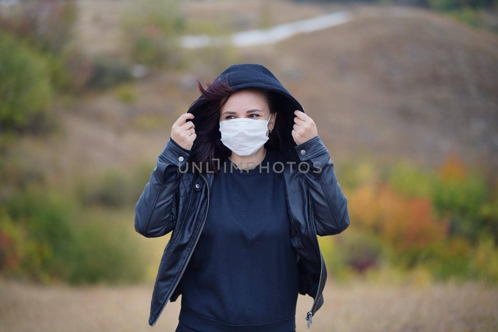 Young woman in medical mask standing in countryside. Adult brunette in protective mask in cool weather on walk in hilly terrain. by epidemiks