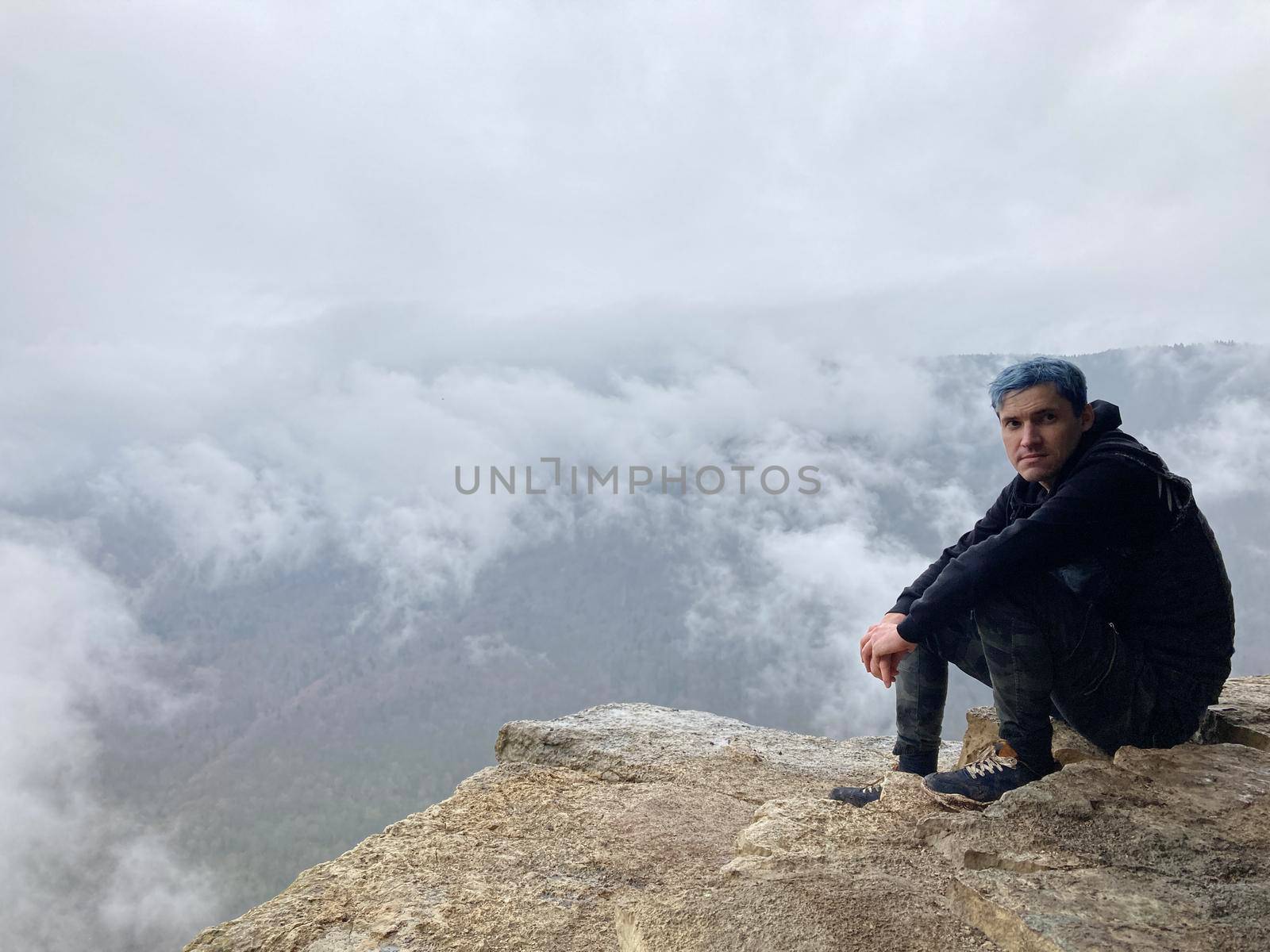 Young man with blue hair sitting on edge of cliff in foggy and cloudy weather. Male tourist enjoys spectacular view of nature, sitting on high rock