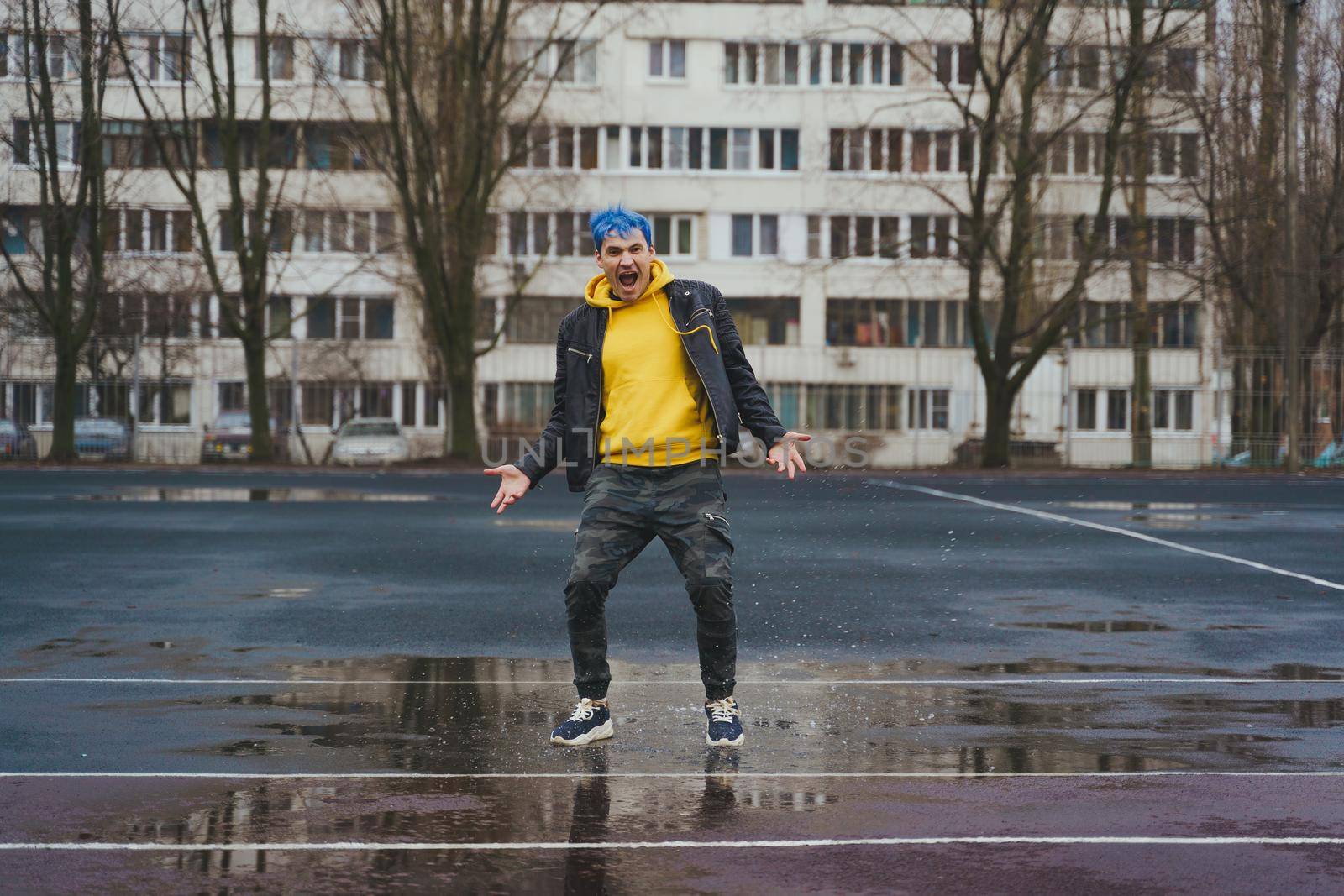 Young man standing in puddle on sports stadium on background of high-rise building. Happy guy with blue hair having fun on city street in springtime. by epidemiks